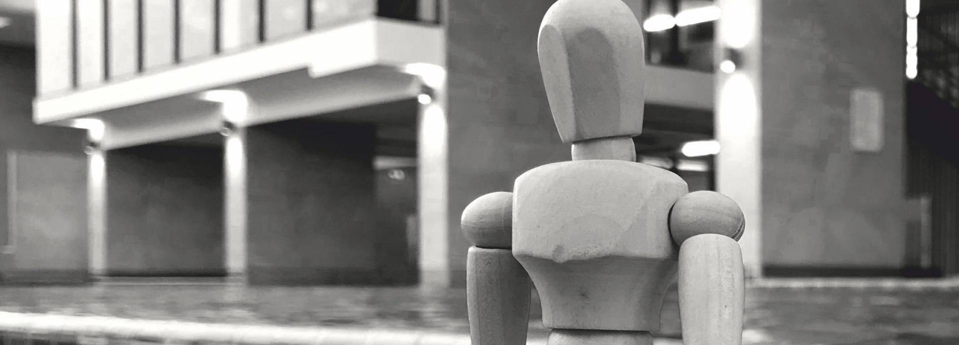 Wooden artist mannequin positioned sitting in front of a concrete brutalist building