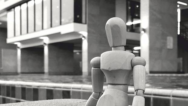 Wooden artist mannequin positioned sitting in front of a concrete brutalist building