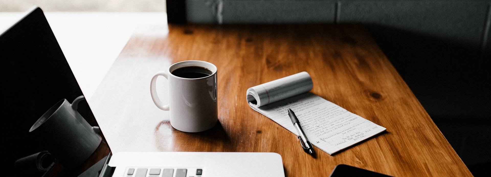 A MacBook, iPhone, flip-pad, pen and cup of coffee sit on a dark wood desk near a bright window.