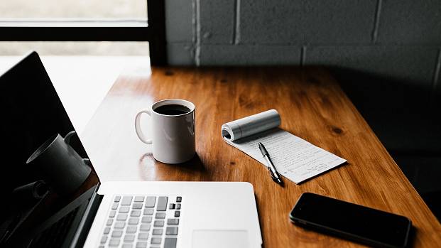 A MacBook, iPhone, flip-pad, pen and cup of coffee sit on a dark wood desk near a bright window.