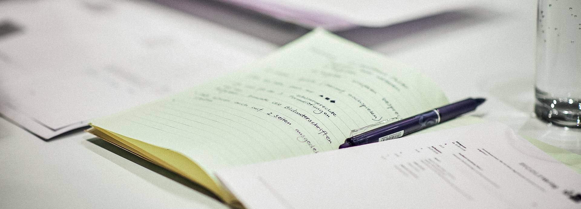 A book full of notes lays open on a table alongside other stacks of paper and a pen.
