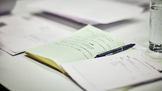 A book full of notes lays open on a table alongside other stacks of paper and a pen.