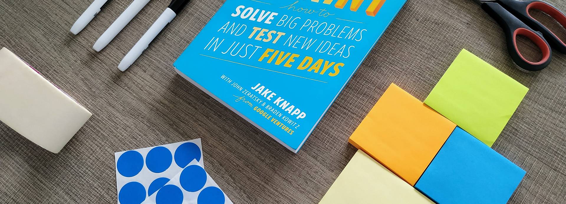 A selection of tools you need for a Design Sprint and How Might We questions, including sticky notes, voting dots, permanent markers and The Sprint Book.