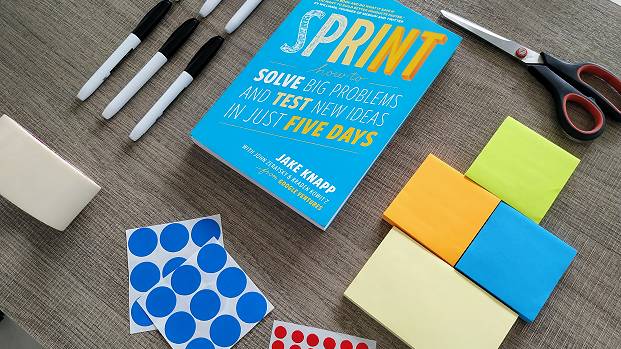 A selection of tools you need for a Design Sprint and How Might We questions, including sticky notes, voting dots, permanent markers and The Sprint Book.