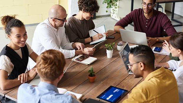 A group of people work together around a desk with a laptop and a tablet.
