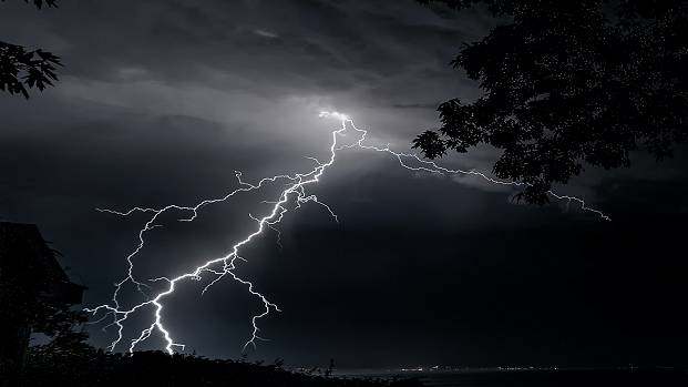 Lightning strikes on a dark night, with trees silhouetted in the foreground.