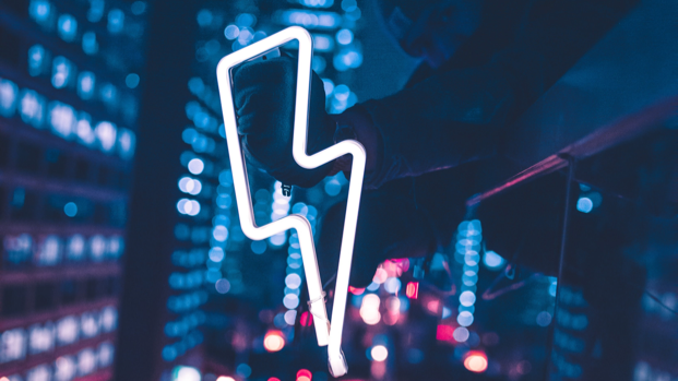 A neon outline of a lightning bolt is held in front of a night-time cityscape.